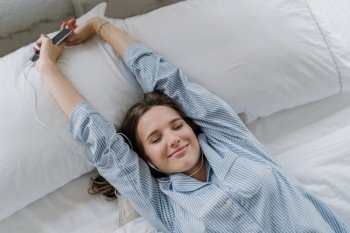 Pleased young European woman stretches in bed, has closed eyes, dressed in striped nightclothes, holds smart phone, enjoys favourite music in earphones, lies on white bedclothes in cozy bedroom