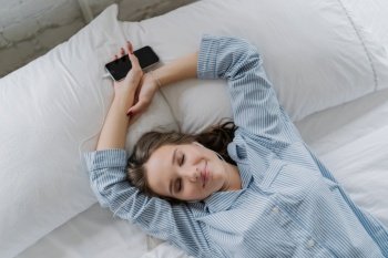 Pleasant looking relaxed young woman has satisfied expression, dreams about something, wears casual clothes, lies on white pillow, connected to smart phone, listens nice music, has rest in bed
