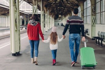 Back view of friendly family hold hands, carry suitcase, going to have voyage trip, pose on railway station platform. Young mother and father, their small daughter wait for training and departure