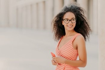 Pleased millennial girl with curly hair, wears pink dress, optical glasses, focused aside with smile, stands outdoor, waits for call, enjoys high speed internet connection, updates new application