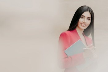 Horizontal shot of lovely brunette woman with happy facial expression, dressed in formal clothes, holds notebook and pen, has black straight hair, stands over white blurred background with free space