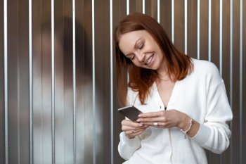 Positive successful European female economist makes money payment on website, poses with cell phone, dressed in white outfit, smiles happily, sends text message, enjoys online communication.