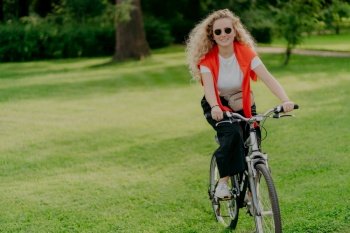 People, nature, rest, lifestyle concept. Happy curly woman rides bicycle among green grass, moves actively, wants to be fit, explores new places in countryside, wears sunglasses, casual clothes