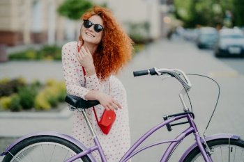 People, leisure, lifestyle and spare time concept. Cheerful curly woman focused into distance, rides bicycle in urban setting, enjoys her hobby, wears shades and long dress, has red small bag