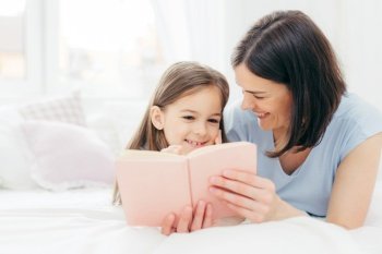 Indoor shot of pleasant looking female child with curious expression, reads interesting book together with her affectionate mother, views colourful pictures, lie on bed in spacious bedroom. Childhood