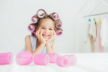 Portrait of cheerful female kid keeps hands under chin, has curlers on hair, going to have nice hairstyle, poses against white background, has charming smile, being in good mood. Children and beauty