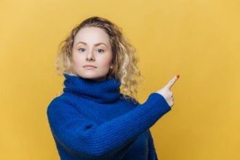 Check this out. Serious blonde young woman with curly blonde hair dressed in bright blue sweater, indicates at blank copy space for your promotional text or advertizing content. Blank yellow wall
