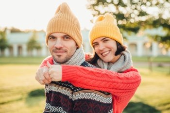 Smiling glad young female in hat and warm cotton sweater embraces her husband who stands back, enjoy spending weekends together, have walk in garden or park, admire splendid sunny autumn weather