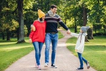 Outdoor portrait of affectionate family walk in park, wear warm knitted clothes. Handsome young man holds daughter`s hand, looks with happy expression at her. Rest and relaxation concept
