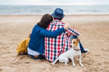 Lovely couple sit at beach near sea, embrace each other have pleasant conversation, being very romantic, admire beautiful nature and their little dog sits near its hosts. People, animals, love concept