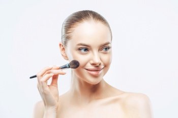 Women, cosmetology concept. Tender healthy European woman gives makeup lesson, applies cosmetic with beauty brush, looks aside with smile, isolated on white background, shows perfect naked body