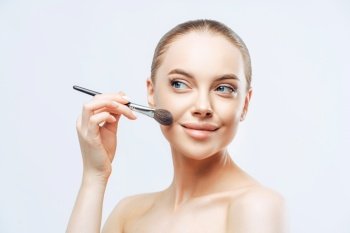 Facial treatment, cosmetology concept. Pretty young woman with dark combed hair, applies powder foundation on face with beauty brush, stands sideways, has naked body, isolated on white background
