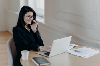 Beautiful female executive makes remote job, talks via mobile phone and checks information in laptop computer poses at workplace surrounded by coffee paper documents notepad has telephone conversation