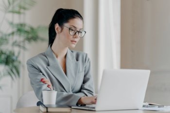 Serious female entrepreneur in eyeglasses, has dark hair combed in pony tail, works on business project, concentrated at laptop computer wears grey formal costume uses information from web resources