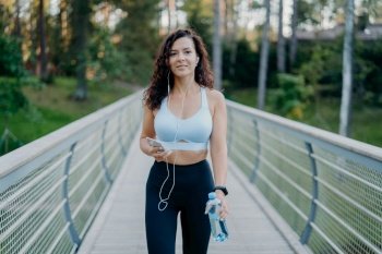 Outdoor shot of slim woman in active wear goes jogging near forest, enjoys listening music in earphones from favorite playlist, holds bottle of water and poses on bridge. Sporty lifestyle concept