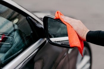 Close up of mans hand wiping water on black car with microfiber cloth. Focus on auto side mirror. Transporation self service