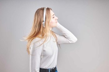 Young woman girl listening to music streaming content having fun watching video enjoying video chat talking with friends making gestures faces using smartphone earphones headphones standing over plain grey background