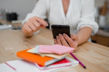 woman using smartphone office supplies technological devices inside home. Young lady using a dark smartphone inside home. Woman holding a black cell phone in a house ambient. Girl with technological devices and office supplies with blank space.