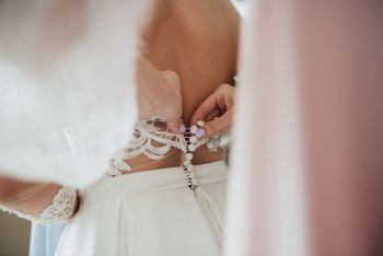 A white wedding dress is knitted to the bride. A bride is being helped to wear the wedding dress. The bride’s preparation for the wedding. Bride white lace wedding dress. Bride help put on the wedding dress