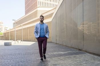 Portrait of a young bearded man, model of fashion, in urban background wearing casual clothes while walking with hands on pocket