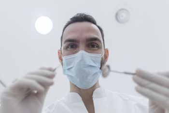 Dentist wearing surgical mask while holding angled mirror and drill, ready to begin