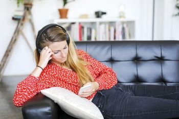 Happy woman listening to music wearing headphones using a smartphone sitting on a sofa in the living room of a interior house