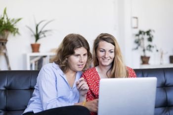 Two young women using computer while sitting on couch in living room