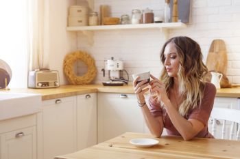 Blonde European female wearing beige dress sitting in bright modern kitchen holding mug in her hands. Picture with selective focus. Quarantine. Self isolation. Stay home concept.