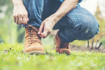 Footwear concept.Handsome man wear jeans knelt down to do up his shoelaces. Preparing before go to party. Brown boots on green grass. close up man hands tie up shoes.