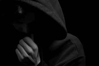  a young guy in a black hoodie on a black background with his face covered.