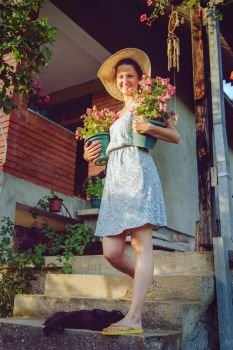 Young woman female farmer happy smiling carry two pots with flowers by her house garden flower pot taking care of the plants wearing hat and summer dress
