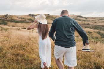 Young couple in love on the mountain range in summer or autumn day walking in nature freedom she is holding his hand and leading wearing hat back view
