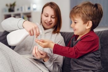 Portrait of small caucasian boy male child kid sitting on the sofa bed at home by his mother young woman spraying alcohol disinfectant antibacterial prevention hygiene product to his hands cleaning