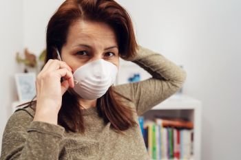 Portrait of mature caucasian woman wearing antivirus antibacterial protective mask during pandemic while standing in front of the bookshelf at work or home preventing disease spread front view