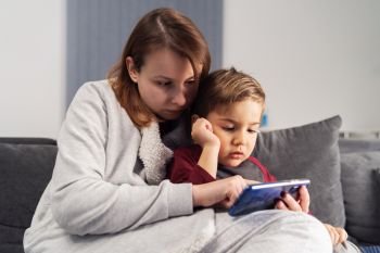 Mother and son young woman and her boy child caucasian kid sitting on the sofa bed at home making video call or playing video games on mobile smart phone during quarantine lockdown