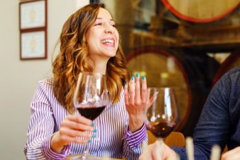 Young caucasian woman beautiful girl brunette sitting by the table at home or restaurant holding a glass of red wine talking and gesturing with hand smiling happy wearing shirt explaining