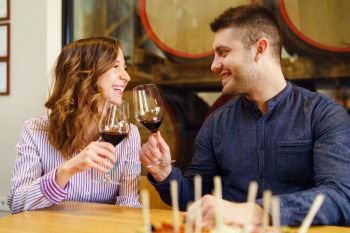 Young beautiful caucasian couple in love toasting with glasses of red wine while sitting by the table at home or restaurant man and woman girl looking each other laughing smiling happy having fun