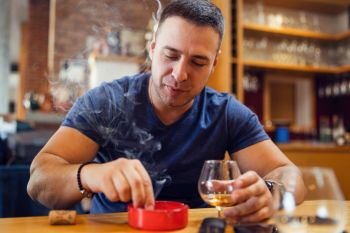 Portrait of young caucasian man adult sitting by table extinguishes a cigar smoking looking to ash tray wearing t-shirt blue in restaurant or home holding glass of cognac or brandy