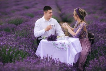 young couple are sitting on chairs at table and drink coffee or tea in lavender field. woman in purple dress and flower’s wreath with man outdoors.. young couple are sitting on chairs at table and drink coffee or tea in lavender field. woman in purple dress and flower’s wreath with man outdoors