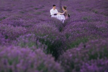 young couple are sitting on chairs at table and relaxing in lavender field. woman in purple dress and flower’s wreath with man outdoors.. young couple are sitting on chairs at table and relaxing in lavender field. woman in purple dress and flower’s wreath with man outdoors