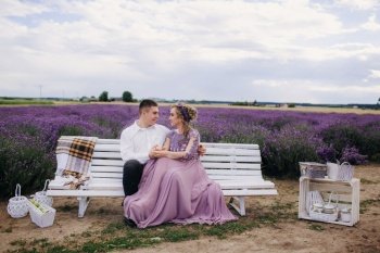 young beautiful couple sitting on a white bench in a lavender field. man and woman in purple dress are hugging outdoors. love story.. young beautiful couple sitting on a white bench in a lavender field. man and woman in purple dress are hugging outdoors. love story