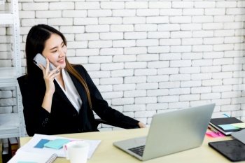 Happy of successful asian young businesswoman hold pick up smartphone working with laptop computer in on White wooden table in white wall office background