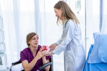 Happy Female doctor therapeutic advising with positive emotions holding exchanging gifts and Give a present to a man patient sit in a wheelchair in hospital background.