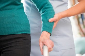 Senior Woman’s Hands On Walking Frame With Care Worker In Background