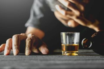Alcohol in a small glass is placed on a black table while a man is sitting in a drunken state.