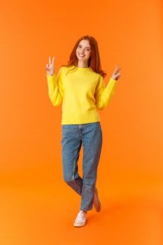 Vertical full-length cute lovely redhead teenage girl in jeans and winter yellow sweater standing with peace signs over orange background, smiling posing express carefree joy emotions.. Vertical full-length cute lovely redhead teenage girl in jeans and winter yellow sweater standing with peace signs over orange background, smiling posing express carefree joy emotions