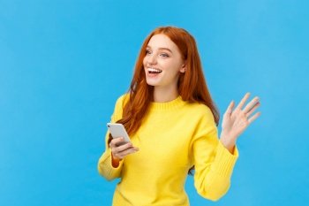 Cute and friendly, outgoing woman with red hair holding smartphone, messaging and waving at someone to turn attention, saying hi, smiling with hello, standing blue background. Copy space. Cute and friendly, outgoing woman with red hair holding smartphone, messaging and waving at someone to turn attention, saying hi, smiling with hello, standing blue background