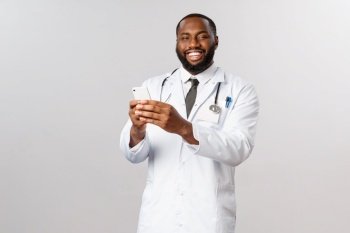 Covid19, pandemic and online medicine concept. Happy friendly african-american doctor using mobile phone and smiling pleased, hold smartphone, receive appointment info in application.. Covid19, pandemic and online medicine concept. Happy friendly african-american doctor using mobile phone and smiling pleased, hold smartphone, receive appointment info in application