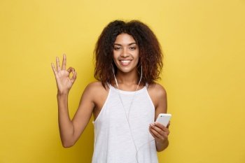 Lifestyle Concept - Portrait of beautiful African American woman joyful listening to music on mobile phone and show ok sign with fingers. Yellow pastel studio background. Copy Space. Lifestyle Concept - Portrait of beautiful African American woman joyful listening to music on mobile phone and show ok sign with fingers. Yellow pastel studio background. Copy Space.