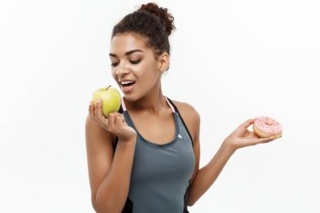 Healthy and diet concept - Beautiful sporty African American make a decision between donut and green apple. Isolated on white background. Healthy and diet concept - Beautiful sporty African American make a decision between donut and green apple. Isolated on white background.